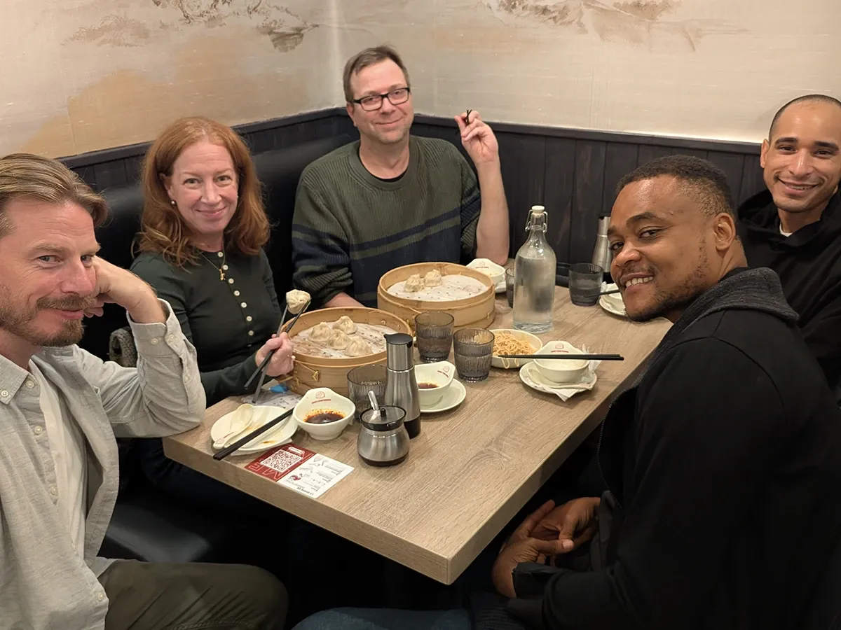 Five teammates smiling around a booth table with bamboo steamers of dumplings, one of them holding a dumpling with chopsticks.
