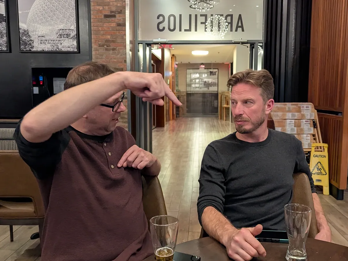 A teammate telling a story with a raised hand at a hotel bar table while another listens, with beer glasses in front of them.