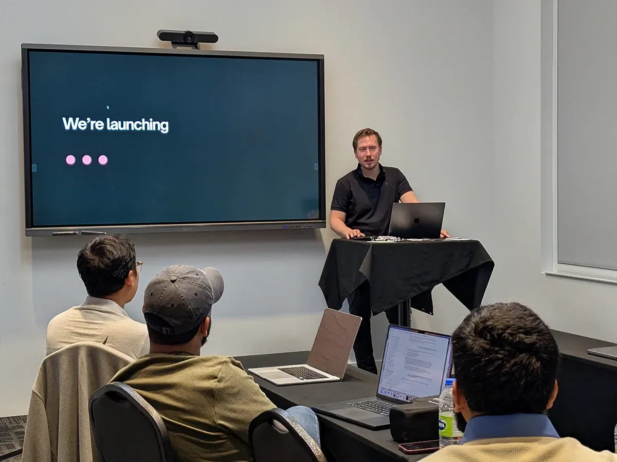Agustin Barrientos presenting from a standing table next to a large screen that says We're launching, with teammates watching from the tables in front of him.