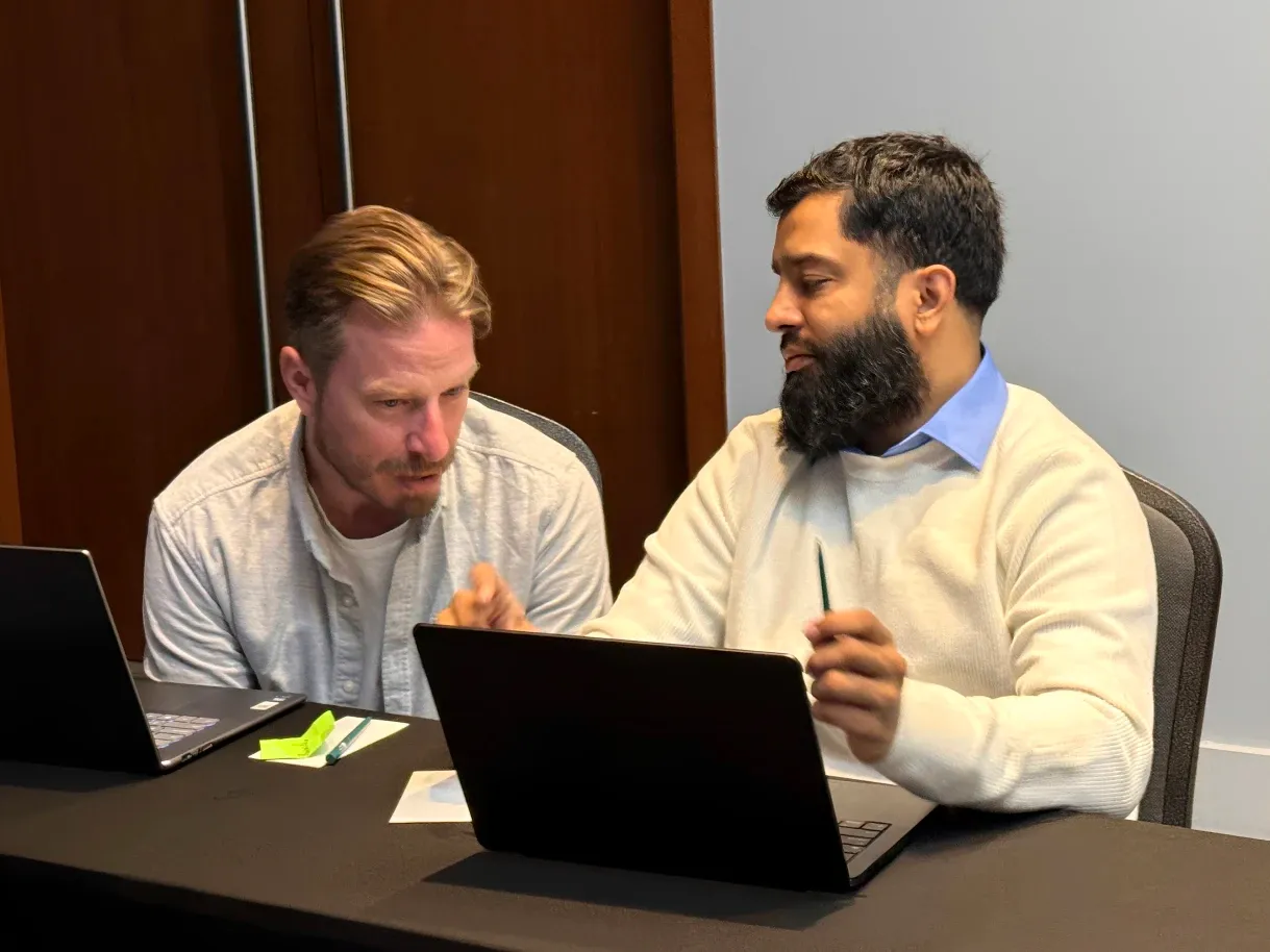 David Chambo and Muneeb Khatri sharing a table with their laptops, one leaning in while the other explains something with a pen in his hand.