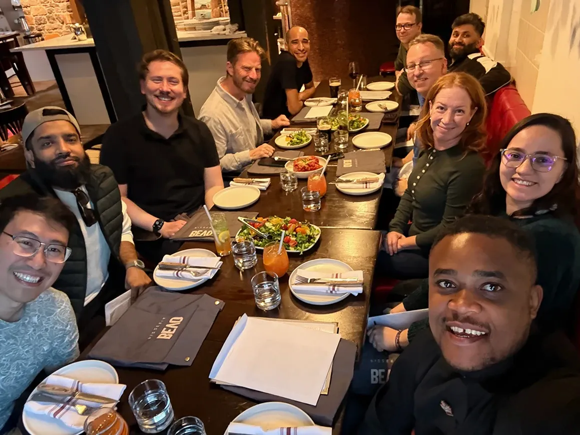 A selfie down a long restaurant table at Bevo Pizzeria in Montreal, with eleven people smiling at the camera from both sides, and salads, drinks, and folded aprons on the table. Ken Young, Ryan Moore, Agustin Barrientos, Daniela Rodriguez Daza, Saad Siddique, Muneeb Khatri, David Chambo, Curtis Krauter, Brendan Da Costa, Kimberly Campbell, and Aniefiok Ntia are visible.