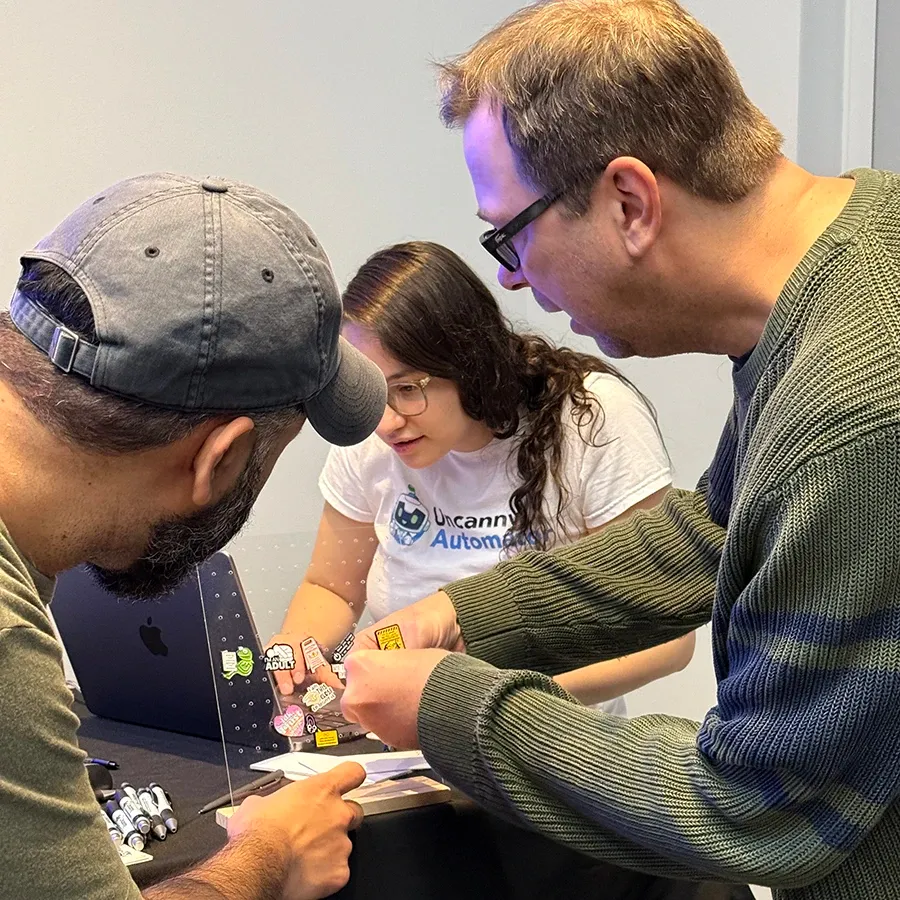 Curtis Krauter and Saad Siddique leaning over the prize table to pick pins from a clear display, while Daniela Rodriguez Daza looks on.