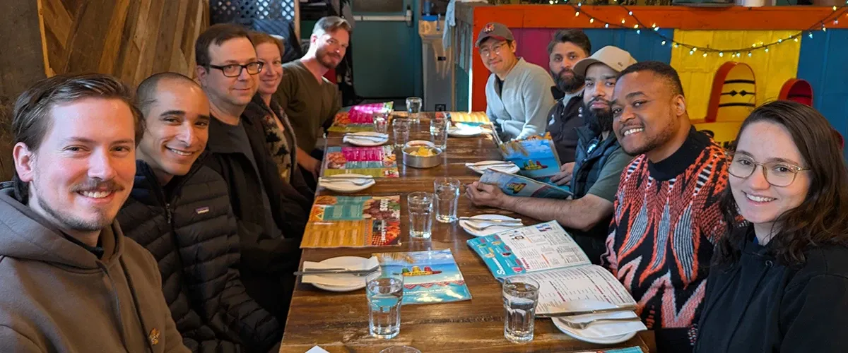 Ken Young, Agustin Barrientos, Daniela Rodriguez Daza, Saad Siddique, Muneeb Khatri, David Chambo, Curtis Krauter, Brendan Da Costa, Kimberly Campbell, and Aniefiok Ntia seated down both sides of a long wooden table in a colorful Mexican restaurant, looking at the camera.