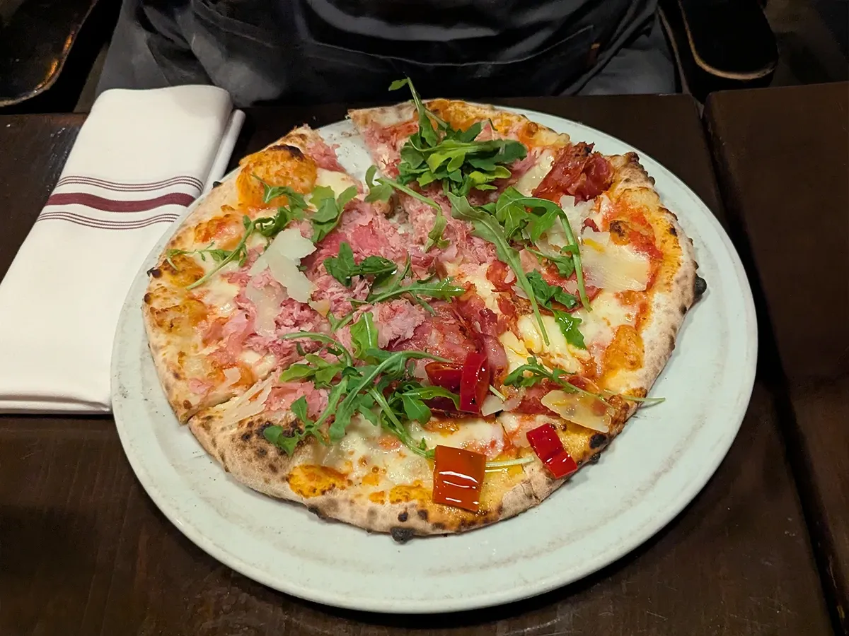 A pizza with ham, salami, roasted peppers, shaved cheese, and arugula on a white plate, with a striped napkin beside it.