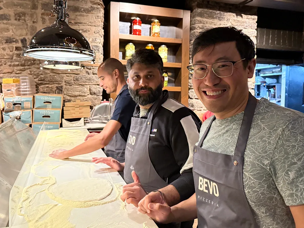Ken Young, Saad Siddique and Brendan Da Costa stretching pizza dough at the counter, Saad is smiling and giving a thumbs up.