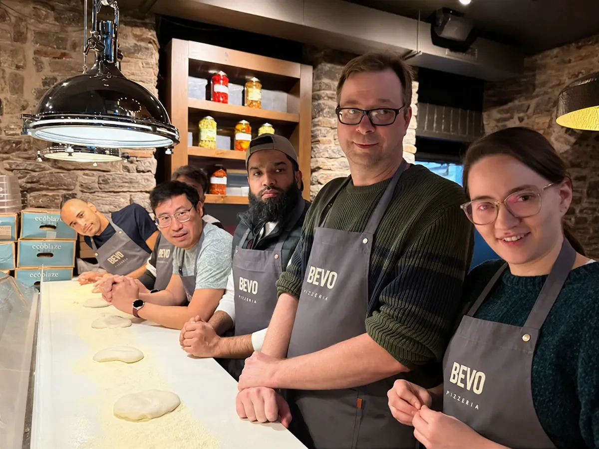Daniela Rodriguez Daza, Curtis Krauter, Muneeb Khatri, Ken Young, Saad Siddique and Brendan Da Costa teammates in gray pizzeria aprons lined up along a flour-dusted counter with rounds of dough, smiling at the camera.