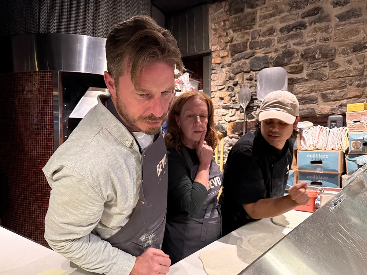 David Chambo and Kimberly Campbell in aprons watching closely as the pizzeria instructor spreads sauce on the dough.